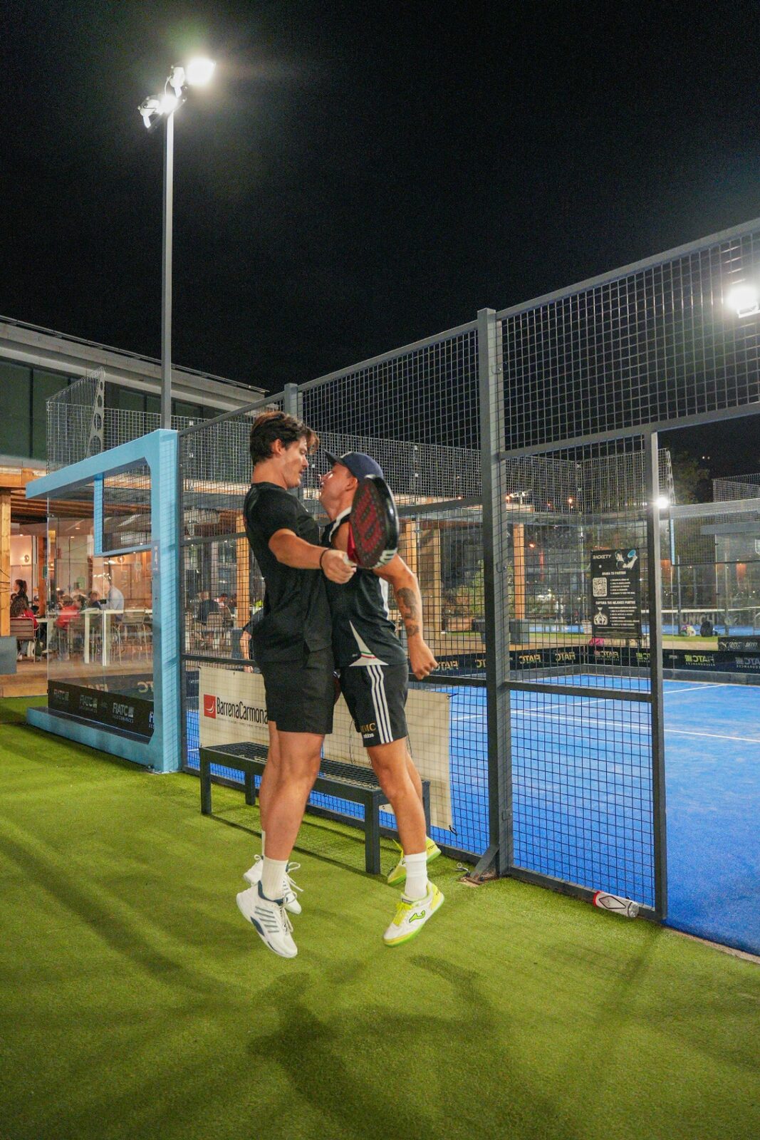Padelspelers vieren een punt onder de lichtmasten tijdens Voyas Padel Nights